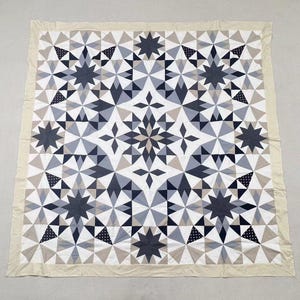 May include: A square quilt featuring a geometric star pattern in shades of charcoal, slate gray, white, and beige. The design incorporates intricate triangular and diamond-shaped fabric pieces, including some sections with a white polka dot print.