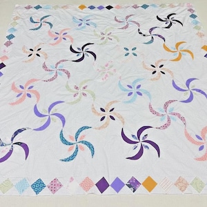 May include: A white quilt with a repeating pattern of colorful pinwheels in shades of purple, pink, blue, and orange. The quilt has a diamond border with various patterned fabrics. The overall design is a traditional patchwork style.