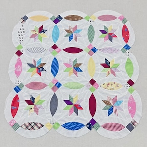 May include: A square quilt featuring a white background with a repeating pattern of circular shapes and eight-pointed stars. The design incorporates various colorful fabric patches in shades of red, blue, yellow, green, and pink with diverse patterns.