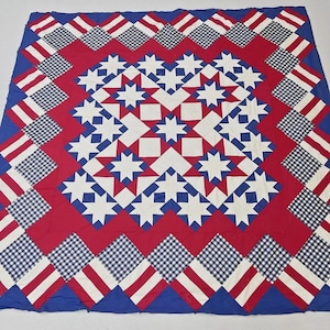 May include: A large, square quilt with a patriotic design. The quilt features a red center with white stars and squares, surrounded by a border of blue, white, and red patterns. The outer border has alternating blue and white checkered squares.