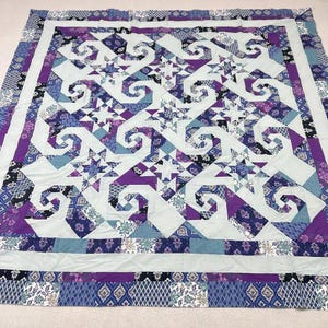 May include: A handmade quilt featuring a complex pattern of swirling designs, stars, and geometric shapes in shades of purple, blue, and white. The quilt has a light blue center and a patterned border.