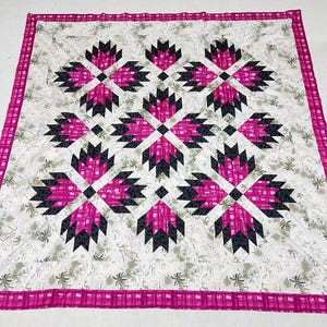 May include: A handmade quilt with a geometric pattern. The quilt features a white background with a repeating design of pink and black shapes. The border is a pink plaid pattern. The quilt is approximately 70 inches by 70 inches.