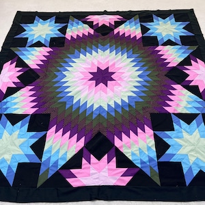 May include: A large quilt featuring a central Lone Star pattern composed of diamond-shaped fabric pieces in shades of pink, purple, blue, and green. The design includes four smaller star motifs in the corners, all set against a black background.