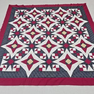 May include: A square quilt featuring a repeating star pattern in shades of deep red, white, and cream, set against a dark gray background with a subtle leaf print. The quilt is finished with a solid deep red border framing the entire design.