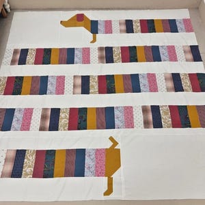 May include: A handmade quilt featuring a dachshund dog design. The dog's body is made of colorful fabric strips in various patterns and colors, including pink, blue, and gold. The quilt is on a white background.