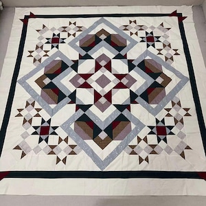May include: A handmade quilt with a geometric design. The quilt features a central pattern of diamonds and squares in shades of red, gray, brown, and white. The border is a dark navy blue with red corner accents. The quilt is made of fabric.