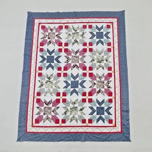 May include: A rectangular quilt featuring a repeating star pattern in shades of pink, blue, and green floral prints on a white background. The quilt is framed by a thin red border and a wide, textured blue border with a scalloped white edge detail.