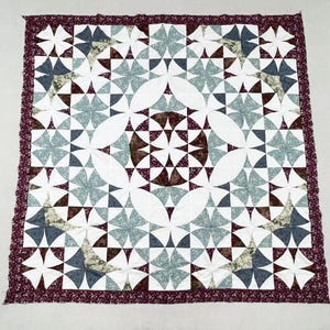May include: A square quilt featuring a complex geometric pattern of white, sage green, navy blue, and dark burgundy floral fabric pieces. The design includes curved shapes and star-like motifs, finished with a wide, dark burgundy floral border.