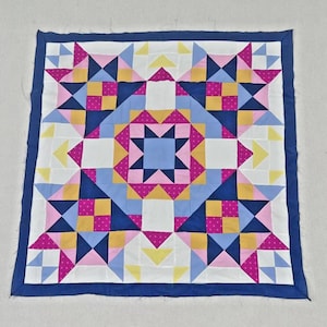May include: A square quilt featuring a geometric star pattern in navy blue, white, light blue, yellow, and magenta pink with white polka dots. The design consists of repeating triangular and square fabric patches bordered by a solid navy blue frame.