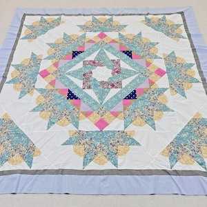 May include: A square quilt featuring a central star pattern composed of white, light blue, yellow, and pink fabric pieces. The design includes floral and polka dot prints, surrounded by a white background and a light blue border with gray trim.