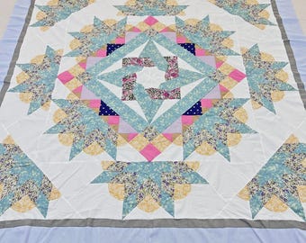 May include: A square quilt featuring a central star pattern composed of white, light blue, yellow, and pink fabric pieces. The design includes floral and polka dot prints, surrounded by a white background and a light blue border with gray trim.