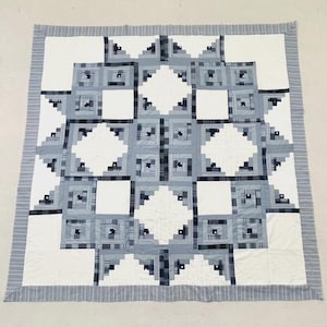 May include: A square quilt featuring a log cabin block pattern in shades of blue, grey, and white. The design consists of alternating solid white squares and intricate pieced blocks with plaid and striped fabric strips, finished with a striped border.