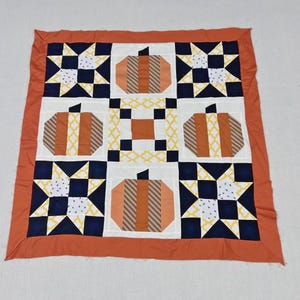 May include: A square quilt featuring a patchwork design with four pumpkin blocks, four star blocks, and a central square block. The quilt is bordered in solid orange fabric and uses a color palette of navy blue, white, yellow, and various shades of orange.