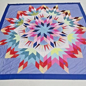 May include: A square quilt featuring a large, multi-pointed star pattern in the center. The design incorporates various fabric colors including pink, blue, red, yellow, and white, with a solid dark blue border framing the entire piece.