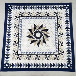 May include: A square quilt featuring a central starburst pattern in navy blue, white, and beige. The design includes concentric borders of pinwheels, triangles, and polka-dot fabric. The outer edge is finished with a solid navy blue fabric border.