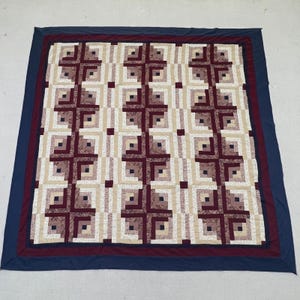 May include: A rectangular quilt featuring a log cabin block pattern in shades of cream, beige, and deep maroon. The quilt is framed by a thin maroon border and a wide navy blue outer border, creating a geometric design across the entire surface.
