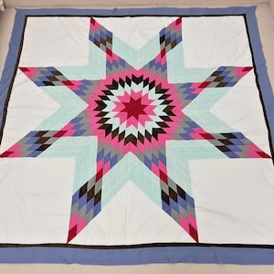 May include: A large, handmade quilt featuring a central eight-pointed star design. The star is composed of diamond-shaped patches in shades of pink, red, gray, brown, and blue, set against a white background. The quilt has a blue border.