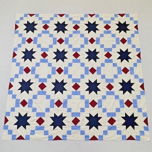 May include: A square quilt featuring a repeating geometric pattern of dark blue eight-pointed stars, light blue square borders, and small red diamond accents set against a cream-colored background. The design is symmetrical and grid-like.