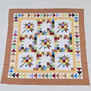 May include: A square quilt featuring a central pattern of five multi-colored leaf motifs made from various fabric squares. The quilt is bordered by a yellow frame and an outer edge of small triangles in diverse colors, finished with a solid brown border.