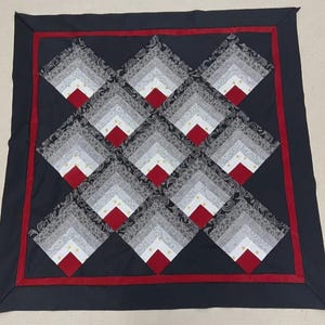 May include: A square quilt with a dark blue border and a red inner border. The quilt's central design features a repeating pattern of diamond shapes in shades of gray, white, and red. The quilt is likely handmade and suitable for a bed or wall hanging.