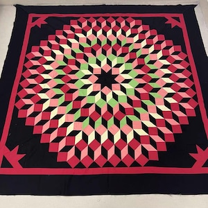 May include: A large, handmade quilt with a geometric design. The quilt features a central starburst pattern in shades of red, pink, and green, set against a black background. A red border frames the design, with corner accents. The quilt is likely made of cotton.