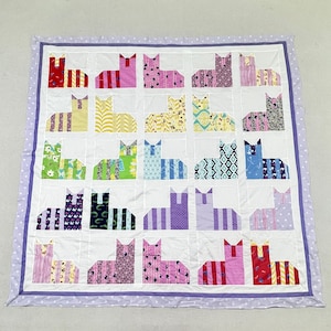 May include: A square quilt featuring a grid of twenty-five cat-shaped fabric patches, each with a unique pattern and color. The quilt is bordered by a light purple frame with white polka dots and a thin dark purple inner trim on a white background.
