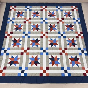 May include: A large, handmade quilt with a repeating star pattern. The quilt features a navy blue border and a grid of gray squares, each containing a star design in red, white, and blue. The overall design is symmetrical and geometric.