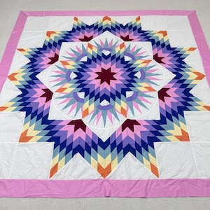 May include: A handmade quilt featuring a large starburst design in shades of purple, blue, pink, orange, and yellow. The star is centered on a white background with a pink border. The quilt is a patchwork design.
