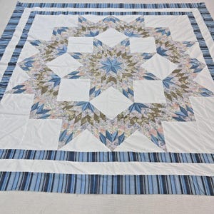 May include: A large, handmade quilt with a central star design. The star is composed of white, blue, and floral patterned fabric. The quilt has a white background and a blue striped border. The overall design is symmetrical and visually appealing.