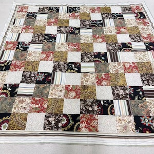 May include: A patchwork quilt with a variety of patterned squares in shades of red, brown, black, and cream. The quilt features floral, paisley, and striped designs. The border is white with thin brown and blue stripes.