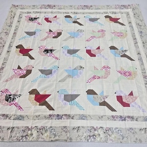 May include: A cream-colored quilt featuring a repeating pattern of stylized birds. Each bird is composed of various patterned fabrics in shades of brown, pink, blue, and gray. The quilt is bordered with a floral print.