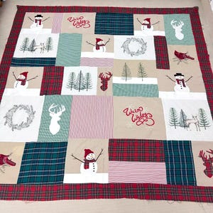 May include: A patchwork quilt with a Christmas theme. The quilt is made of various fabrics in red, green, white, and brown. It features embroidered images of snowmen, reindeer, cardinals, and trees. The text "Warm Wishes" is embroidered in red on a beige fabric square.