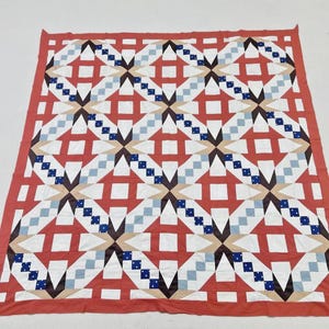May include: A square quilt featuring a geometric pattern of interlocking stars and squares. The design incorporates a color palette of coral red, white, light blue, dark blue with white dots, and tan, all bordered by a solid coral red fabric edge.
