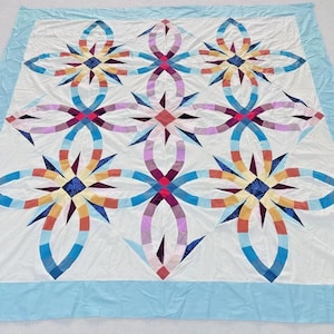 May include: A handmade quilt with a white background and a light blue border. The quilt features a repeating pattern of colorful, interlocking floral designs in shades of blue, red, orange, and purple. The quilt is a large size.