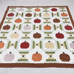 May include: A patchwork quilt featuring a repeating pattern of stylized pumpkins in shades of orange, red, and brown, separated by green and white striped fabric blocks, all framed by a solid brown border on a white background with small dots.