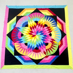 May include: A square quilt featuring a central star pattern composed of vibrant, multi-colored geometric shapes in shades of pink, yellow, blue, green, and red, set against a black background with a bright multi-colored border.