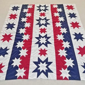 May include: A large, rectangular quilt with a patriotic design. Alternating vertical stripes of red, white, and blue. White stars are sewn onto the red and blue stripes. The quilt is spread out on a light-colored surface.