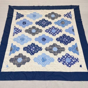 May include: A handmade quilt with a cream-colored center featuring blue and white geometric flower patterns. The flowers have various designs, including polka dots, plaid, and abstract shapes. A dark blue border frames the quilt.