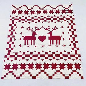 May include: A rectangular quilt featuring a white background with a deep red pattern. The center displays two reindeer facing a central heart. The border consists of a checkered grid and a row of eight-pointed stars in red and white.