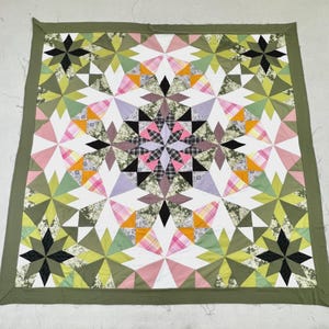 May include: A square quilt featuring a geometric star pattern composed of various fabric patches in shades of olive green, black, white, pink, yellow, and lavender. The design includes plaid, floral, and solid textures with a solid olive green border.