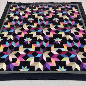 May include: A large, handmade quilt with a geometric starburst pattern. The quilt features a black background with colorful star shapes in shades of pink, purple, blue, yellow, and green. The quilt has a black border.