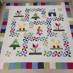 May include: A handmade quilt with a white center featuring colorful appliqued designs of trees, flowers, houses, and animals. The quilt is bordered with a patchwork of various colored fabric strips. The overall design is a charming, folk art style.