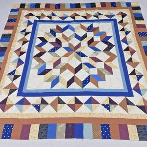 May include: A handmade quilt with a central starburst design in shades of blue, brown, and purple. The quilt features a white background with a blue border and a patchwork border with various patterned fabrics. The quilt is a cozy home decor item.