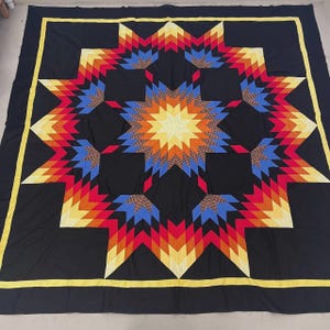 May include: A large, handmade quilt with a striking starburst design. The quilt features a black background with a central star in shades of yellow, orange, and red. The star is surrounded by eight additional stars in complementary colors. A yellow border frames the quilt.