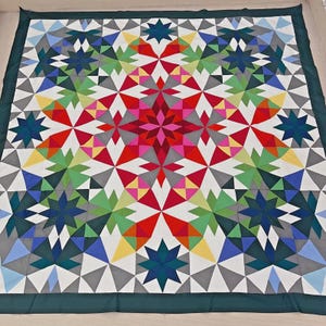 May include: A square quilt featuring a geometric star pattern composed of multi-colored fabric triangles. The design includes shades of red, pink, yellow, light green, dark green, blue, and grey, all bordered by a solid dark green fabric edge.