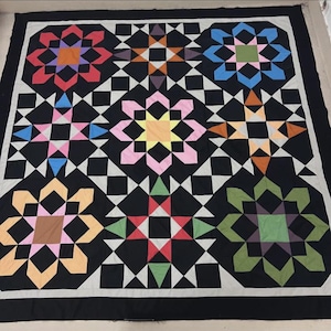May include: A large, handmade quilt with a black background and colorful starburst patterns. The quilt features a variety of colors, including orange, pink, blue, and green, and is bordered with a black and white checkered pattern.