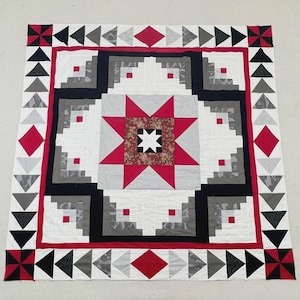 May include: A square quilt featuring a central star pattern in red, white, and black, surrounded by log cabin blocks in shades of gray, white, and black. The border consists of alternating red diamonds and triangles in black, gray, and white.