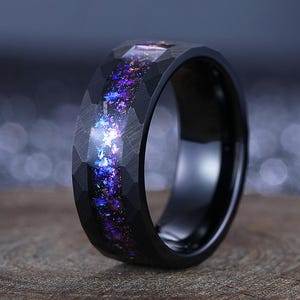 Könnte beinhalten: Ein schwarzer Ring mit einer facettierten Außenband und einem schimmernden Inlay aus Lila, Blau und Weiß. Der Ring liegt auf einer Holzoberfläche und zeigt seine polierte Oberfläche und sein einzigartiges Design.