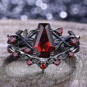May include: A black ring with a large, rectangular, deep red gemstone at the center. The ring features a leaf and vine design with smaller red gemstones. The ring is set on a wooden surface.