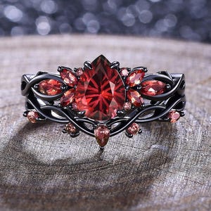 May include: A black ring with a large, pear-shaped, red gemstone at the center, surrounded by smaller red and pink stones. The ring has a detailed, vine-like design. The ring is set on a wooden surface.